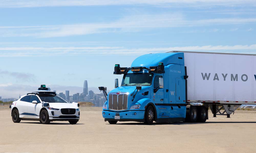 Ein Waymo Taxi und ein Waymo Truck stehen in der W&uuml;ste. Im Hintergrund sieht man die Skyline einer Stadt.